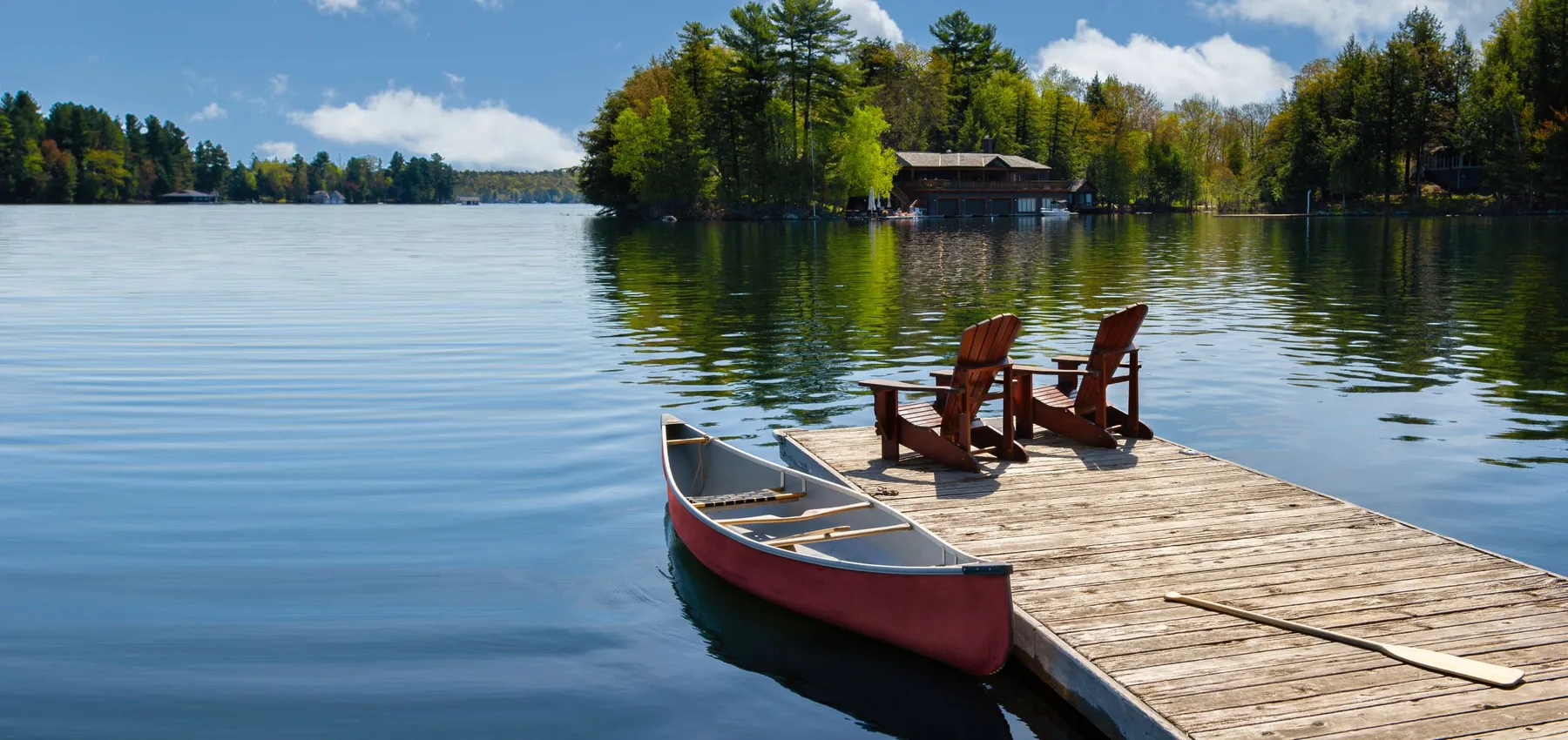 Two Adirondack chairs on a wooden dock overlook a lake. Nearby, a red canoe is tied to the pier, alongside life jackets and a paddle.