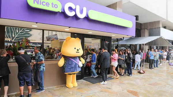 A line of people wait to get into a store. A sign over the door says "Nice to CU" in purple and while lettering and a bear mascot stands outside near the door.