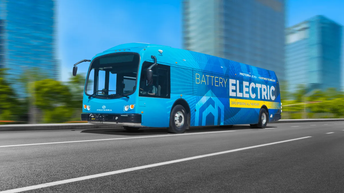 A blue 40-foot transit bus lettered as "Battery Electric" on a multi-lane road with blurred skyscrapers in the background.