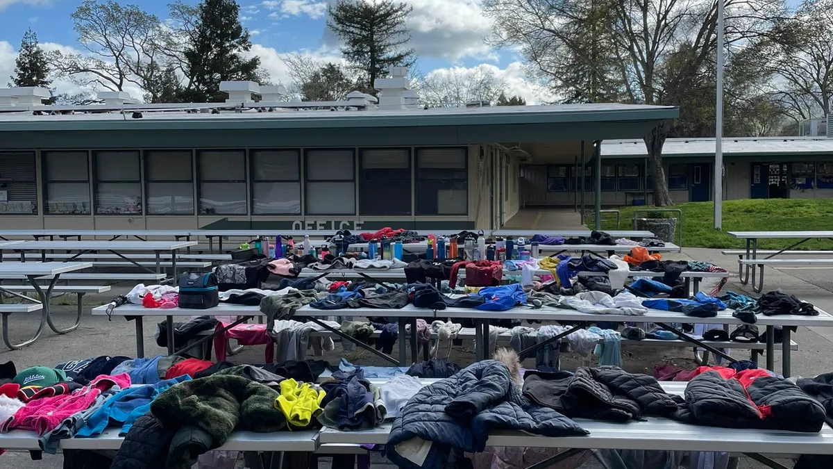 Cafeteria-type tables sit outside of a school building. Clothes are spread out on the tables and seats.