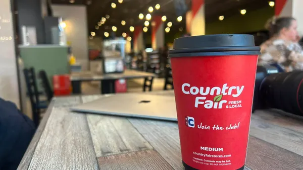 A photo of a cup of coffee on a table inside a store. The side of the cup says "Country Fair Fresh & Local. Join the club. Medium."