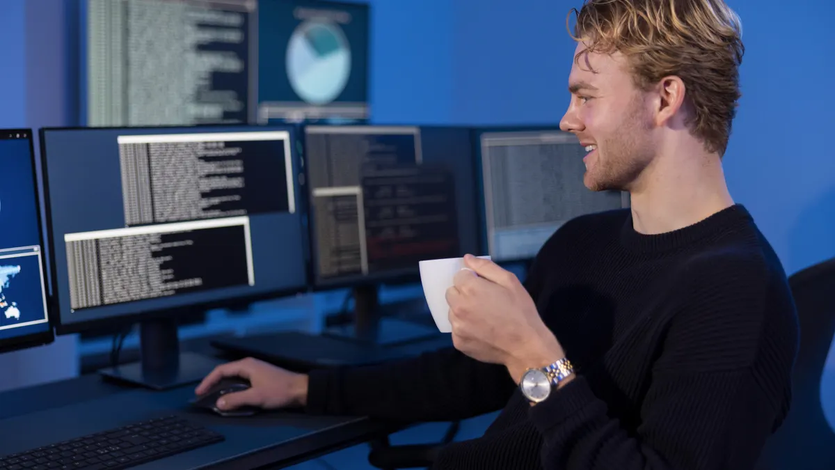 A man holding a cup of coffee sits at a desk that holds several monitors showing data