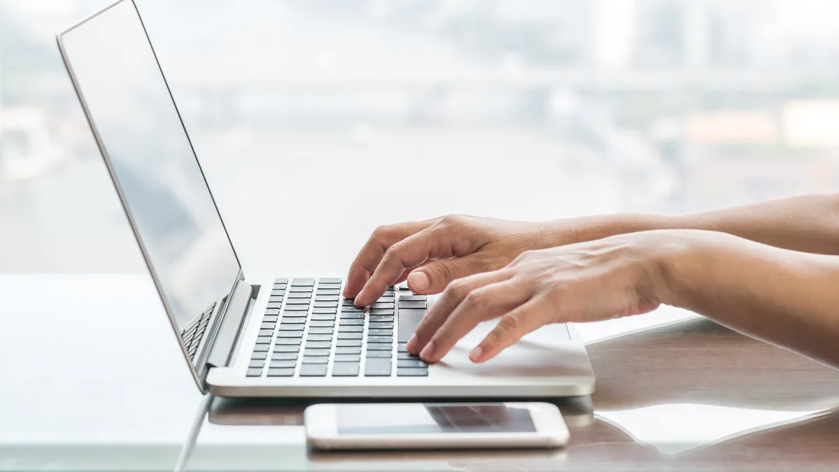 A side view of an open laptop sitting on a table. Hands are on the keyboard. A cellphone sits next to the laptop