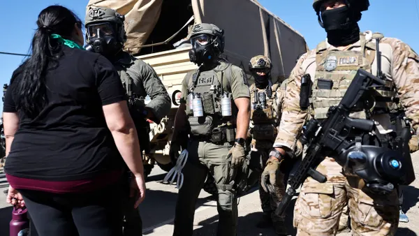 People wearing camouflage suits that say POLICE, helmets and black scarves over their faces, carrying guns, facing a person wearing a black t-shirt and black plants.