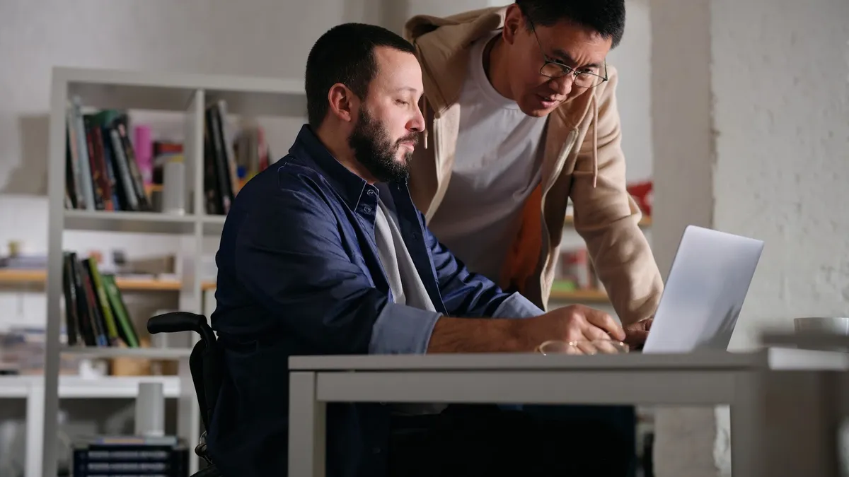 Two people, one in a wheelchair, looking at a laptop at a desk.