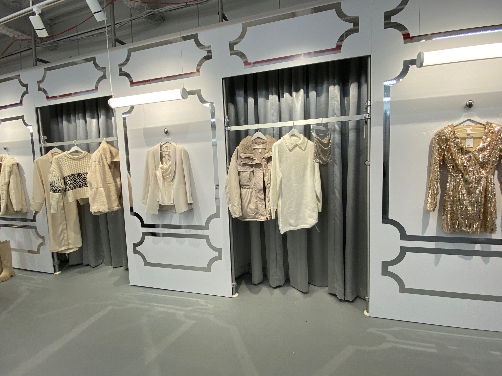 A wall with curtained dressing rooms and a few racks with clothes that are gold and cream colored.