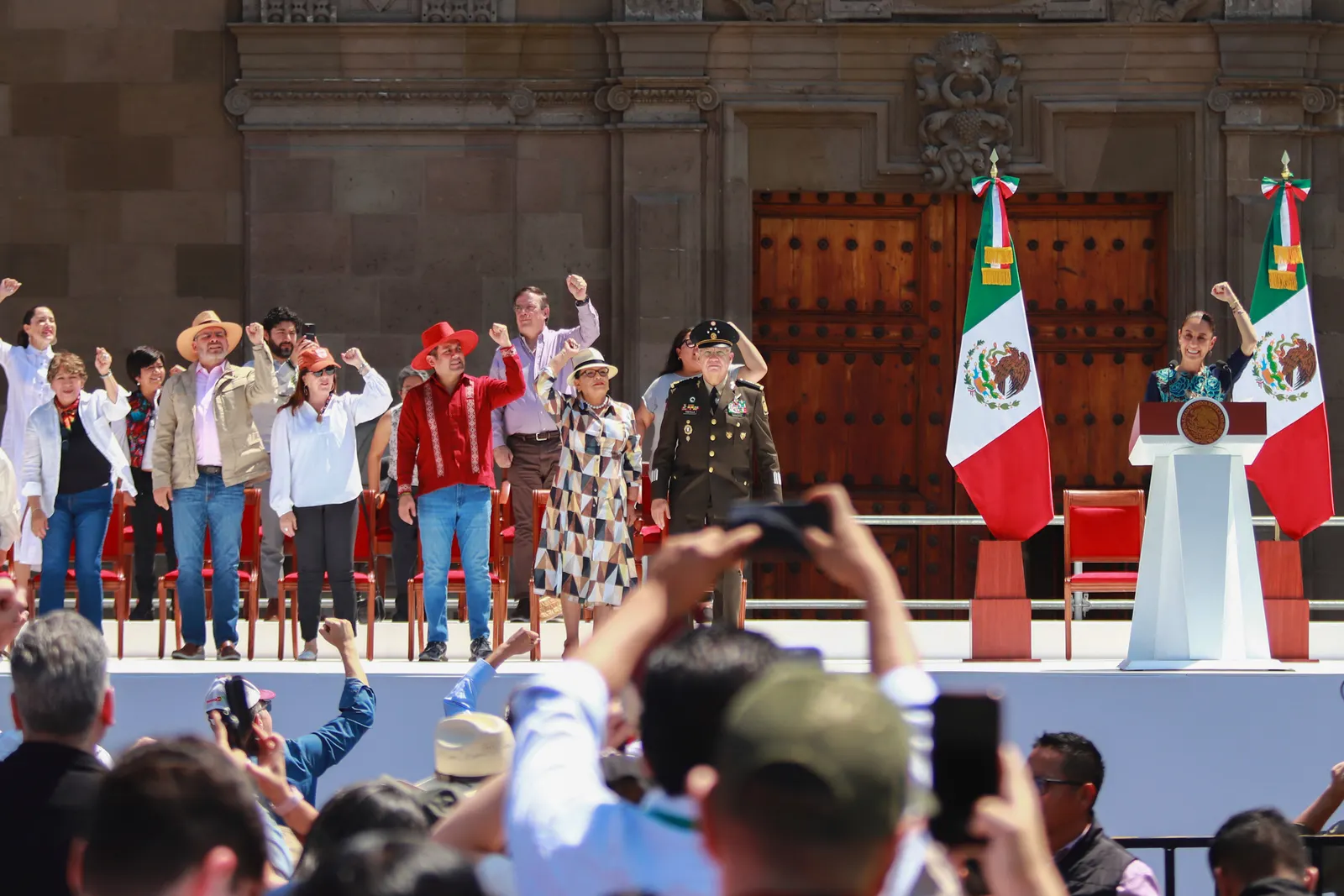 A crowd of people stand  with an arm raised in a fist alongside a pulpit between two Mexico flags. A stone building with big doors is in the background.