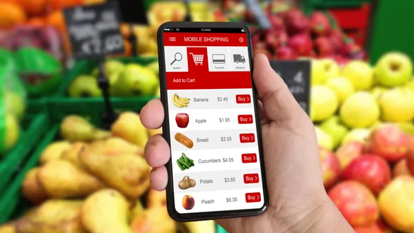 In front of a fruit display, a hand holds a phone with a grocery app open.