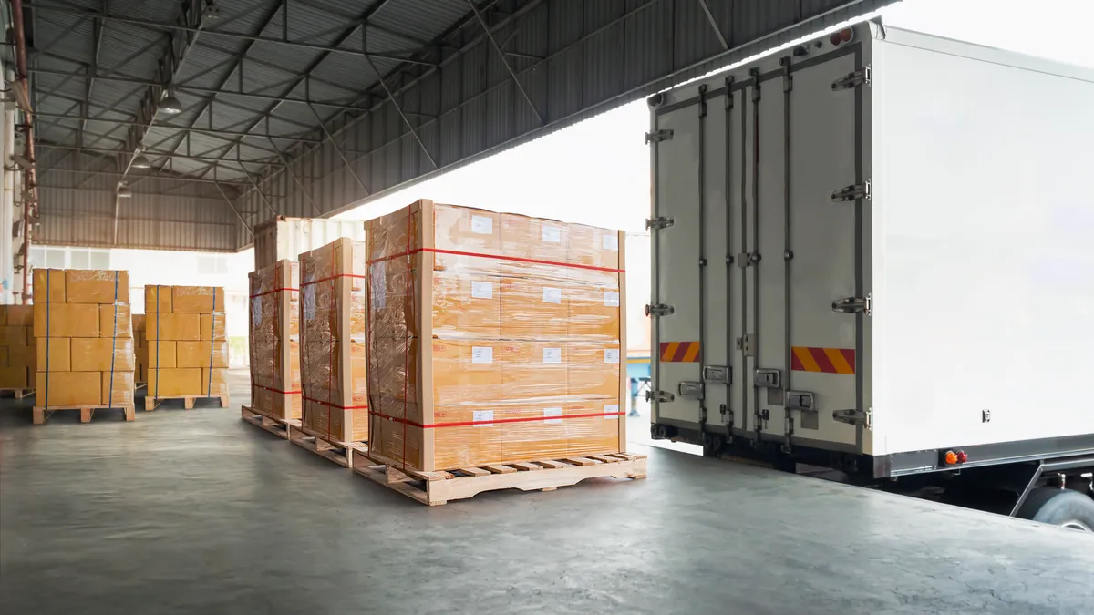 Packaging boxes are wrapped and stacked on pallets in a warehouse, with a trailer at the dock.