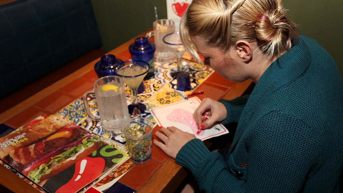 A person with blond hair and braids in a blue sweater colors with a red crayon at a restaurant table, a menu on their left.