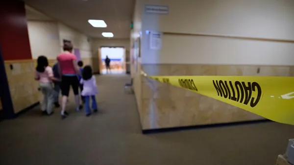 An adult and small children are walking away from the camera down a hallway in a school building. In the forefront is yellow caution tape across another hallway.