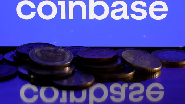 The Coinbase logo is shown on a blue screen. In the foreground, stacks of coins are piled up.