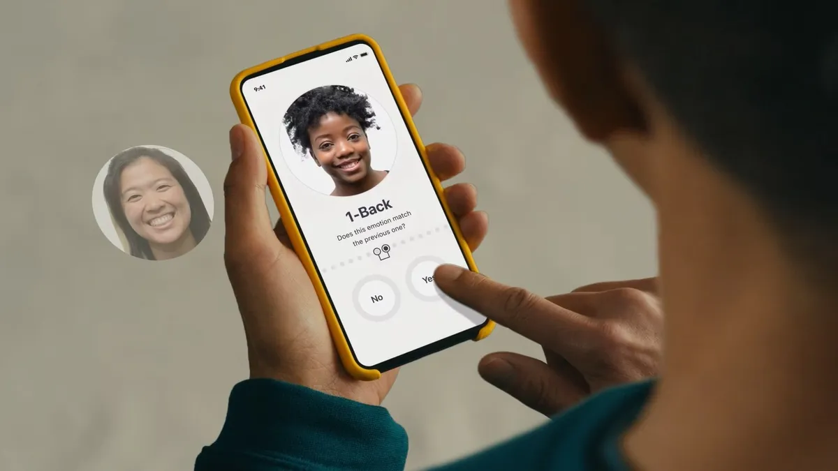 A person holds a phone that shows an image of a face with the prompt: "Does this emotion match the previous one?"