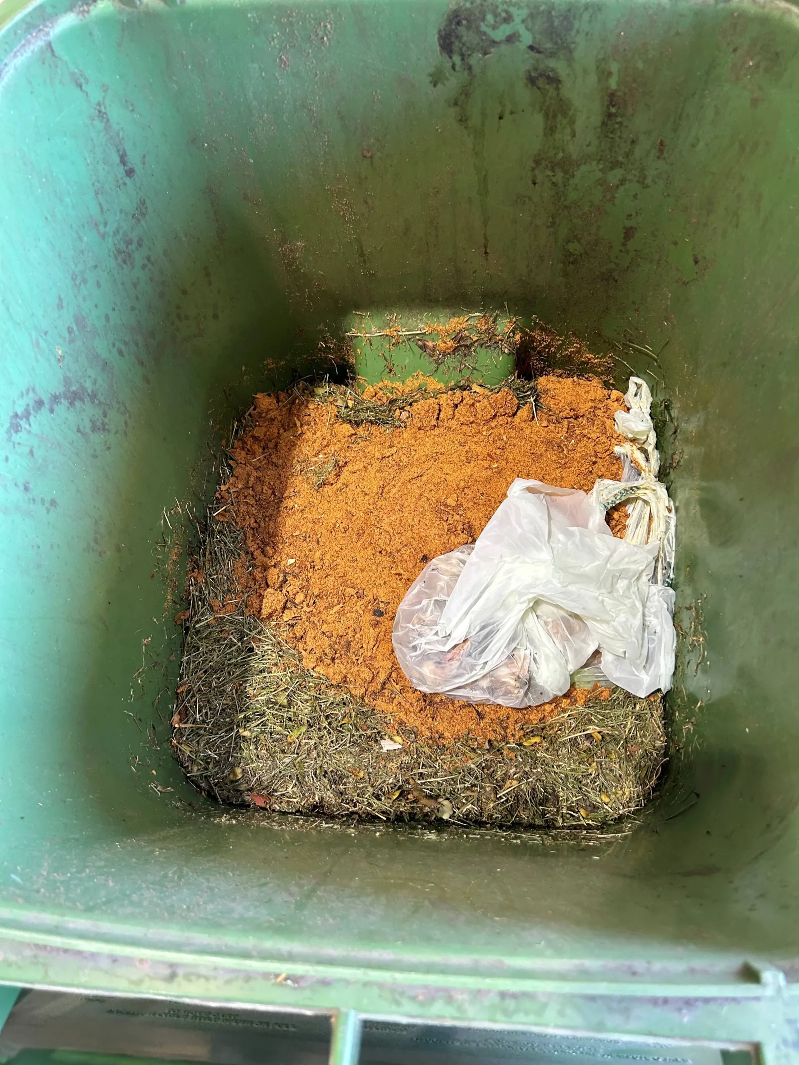 A pile of dried and homogenous-looking organic material lies on top of lawn clippings in a bin, with one bag of additional food scraps.