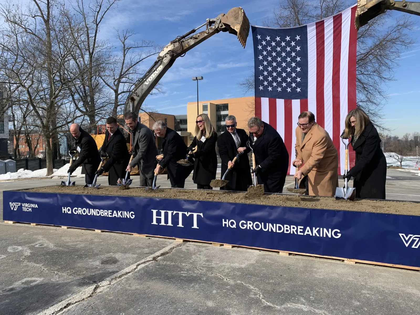 A group of well-dressed people hold up shovels covered in dirt as they stand in front of a box that holds the dirt. An American flag looms behind.