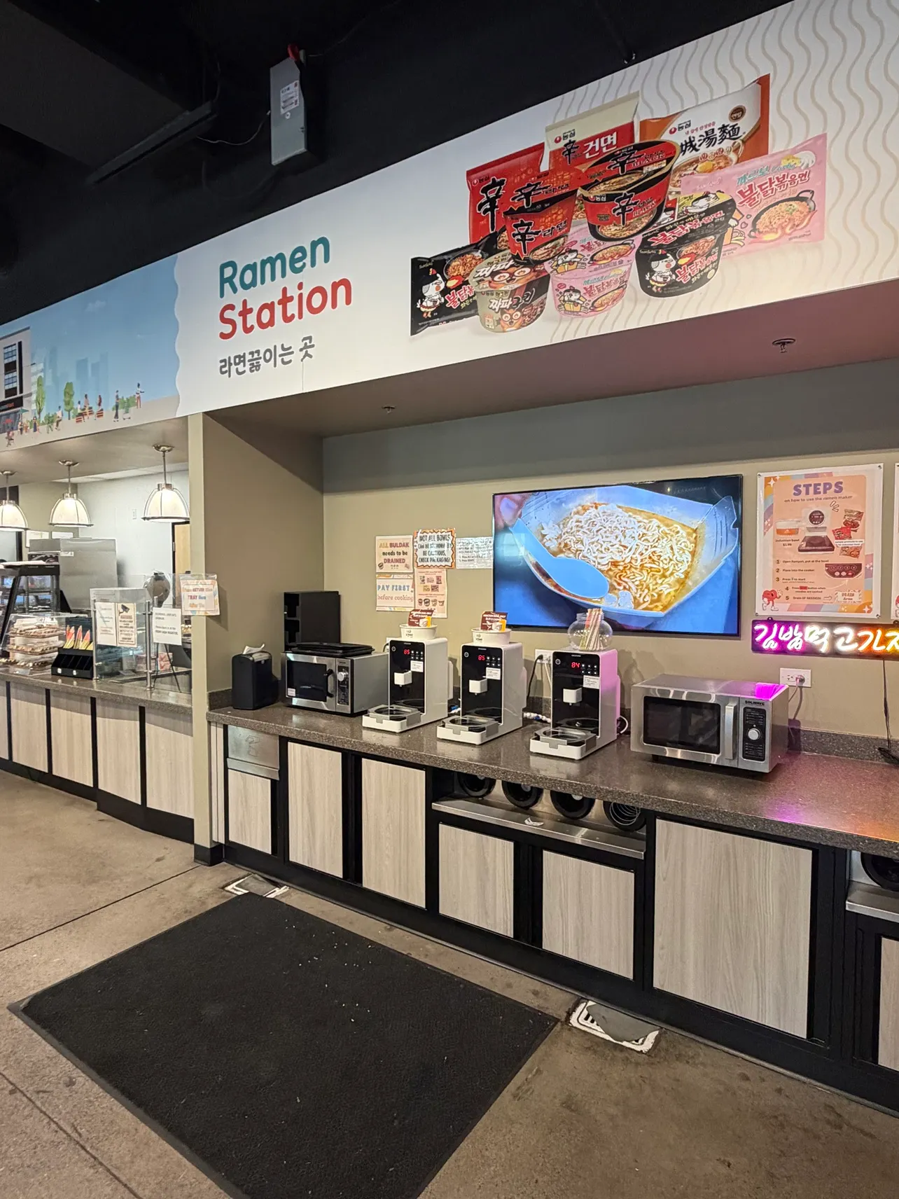The ramen station in a store features devices that dispense hot water as well as microwaves and disposable chopsticks. Signs on the wall notes that all buldak needs to be drained and customers must pay before starting to cook, while a sign over the station reads "Ramen station."