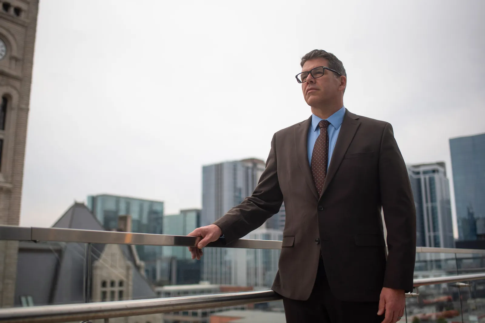 A person in a dark suit and glasses stands on a rooftop balcony with one hand on a railing, looking into the distance with a city skyline and a stone clock tower behind them.