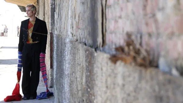A person leans against a stone wall outdoors wearing a top with extra long sleeves that drape on the ground.