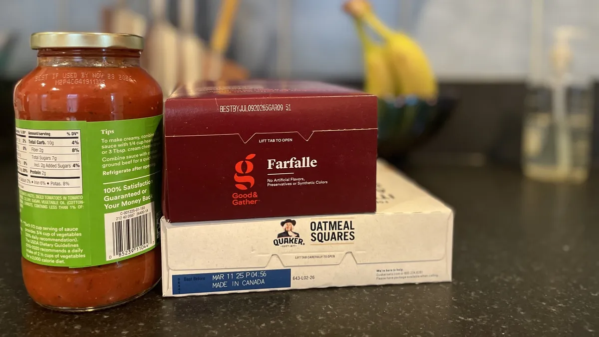 A jar of pasta sauce sits next to boxes of pasta and cereal, with their date labels facing out, on a kitchen counter