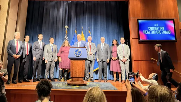 A row of people stand onstage behind a man in a blue suit. A TV screen to their left reads "Combating Healthcare Fraud."