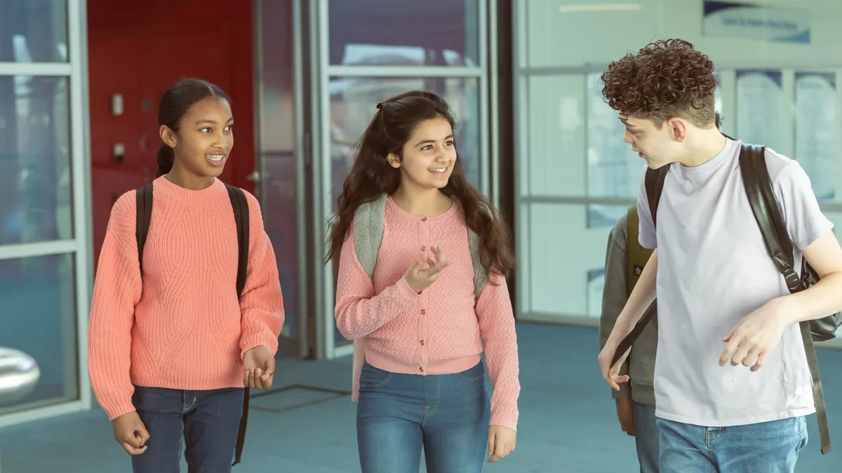 Three middle school students walk in a hallway of a school building. They are looking at each other and carrying backpacks.