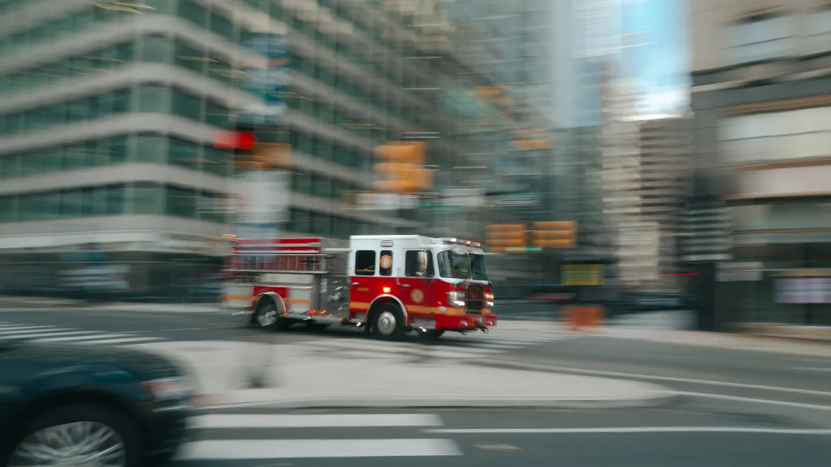 A red fire engine speeds through a city street with tall buildings in the background.