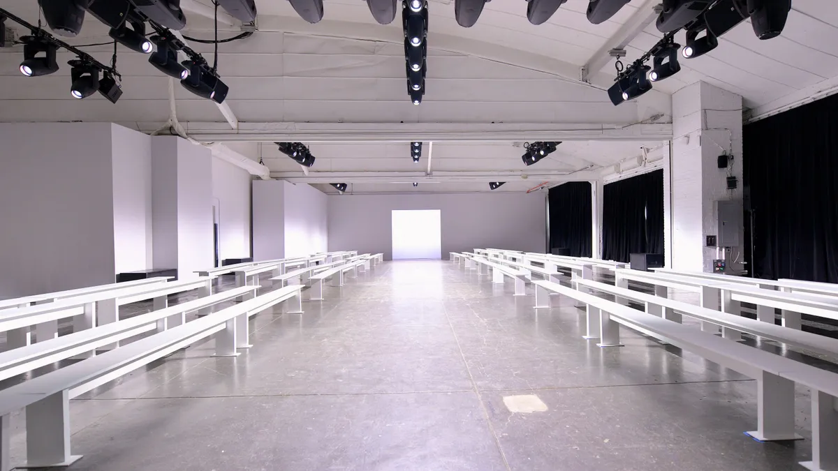 An empty runway in a large white studio is flanked by rows of empty white benches.