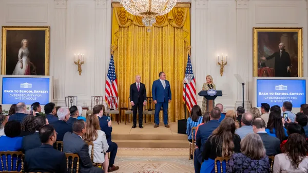 Alejandro Mayorkas, Miguel Cardona and Jill Biden stand in the center of the East Room of the White House between two screens that read “Back to School Safely: Cybersecurity for K-12 Schools.”