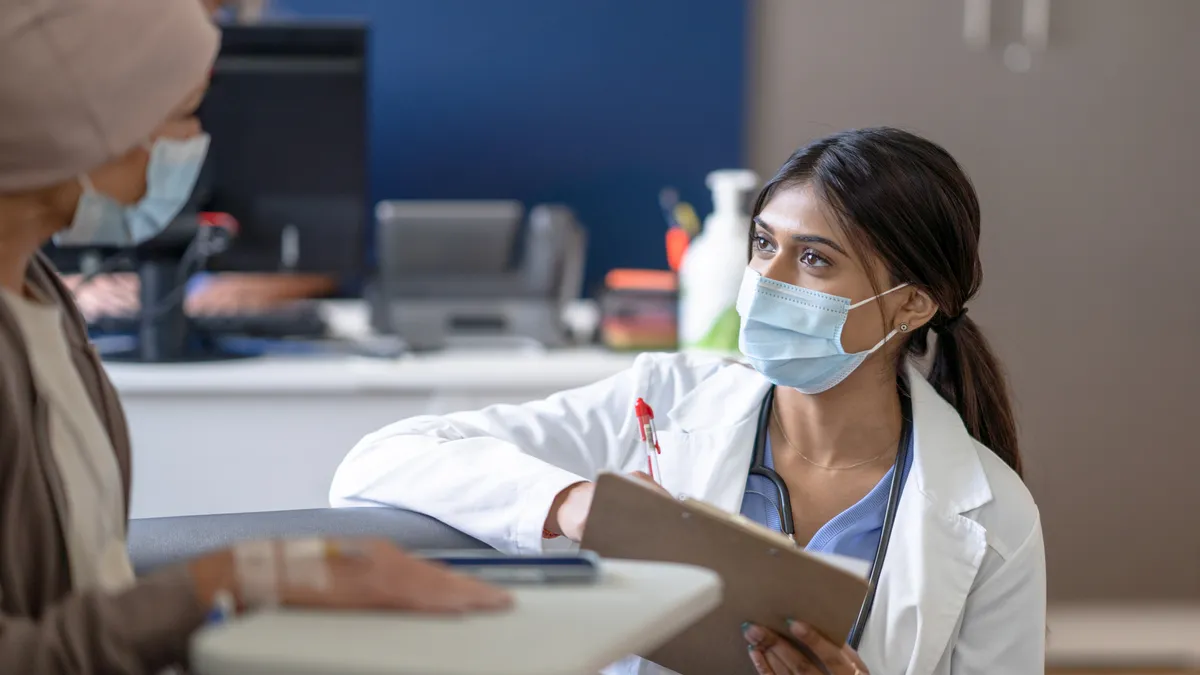 A female doctor bend's down to eye level as she talks with a cancer patient who is receiving her treatment intravenously. She is wearing a white lab coat and has a clipboard in hand as she takes notes