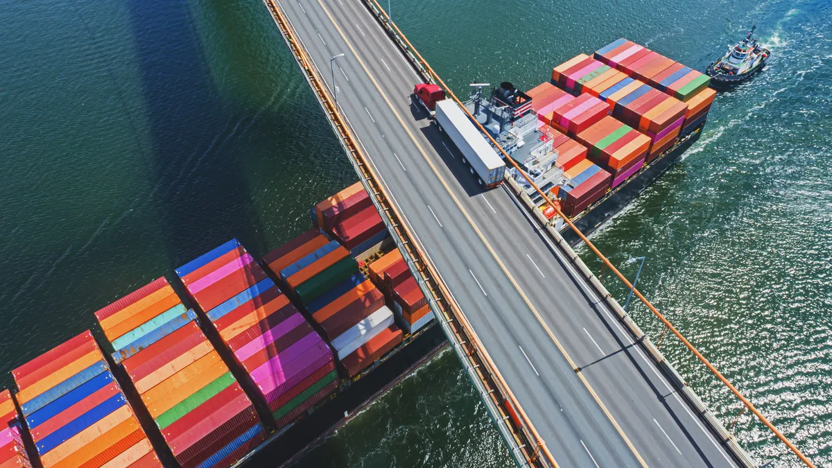 Aerial drone view of a container ship navigating beneath a tractor-trailer crossing a suspension bridge.