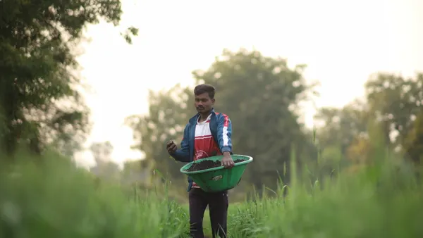 A farmer stands in focus in a green field with biochar in his hand and a bucket of biochar to be spread.