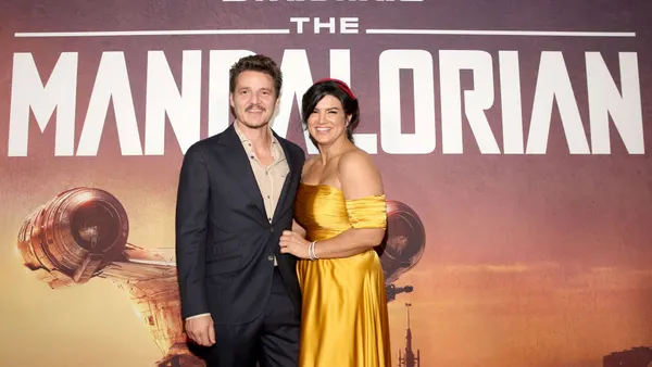 A man and a woman in a yellow dress stand close together on a red carpet in front of a backdrop that says "Star Wars: The Mandalorian."