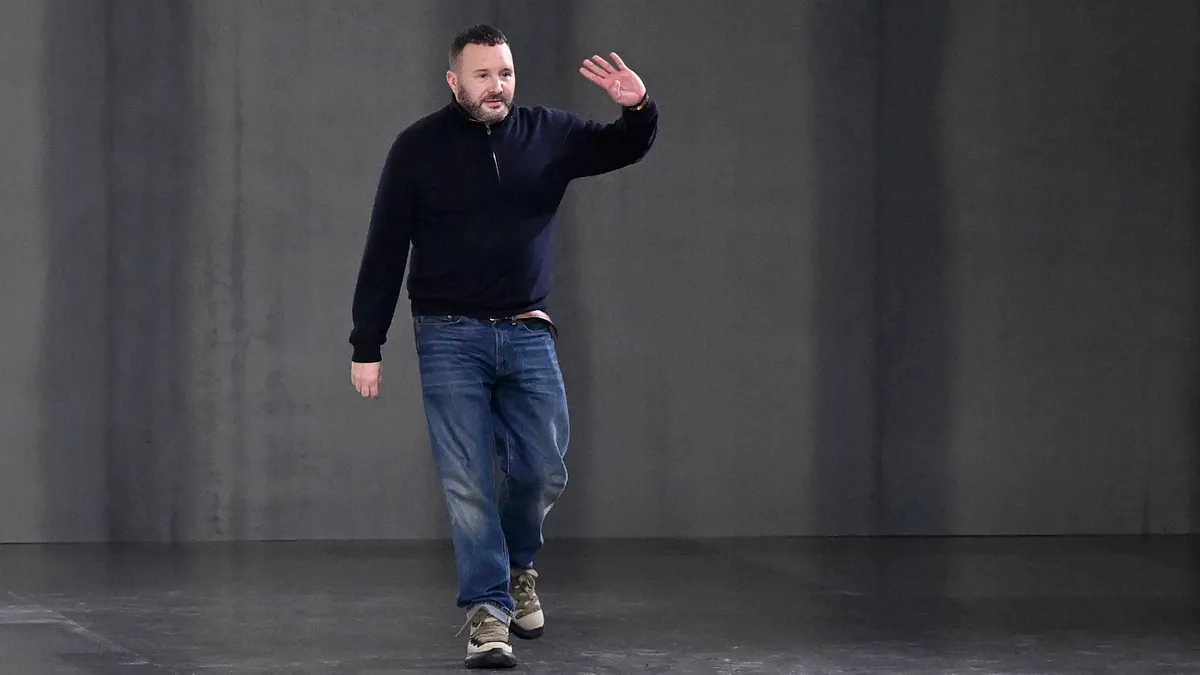 A person wearing blue jeans and a black sweater walks on a fashion runway and waves.