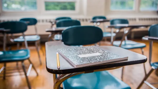A row of desks sit empty in a classroom with the desk in the forefront having a notebook and pencil sitting on top.