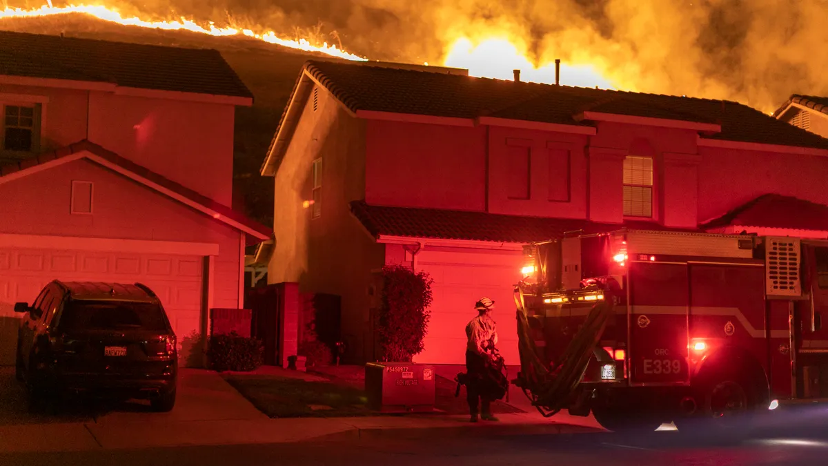 Wildfire burns on a hill above two houses and cars.