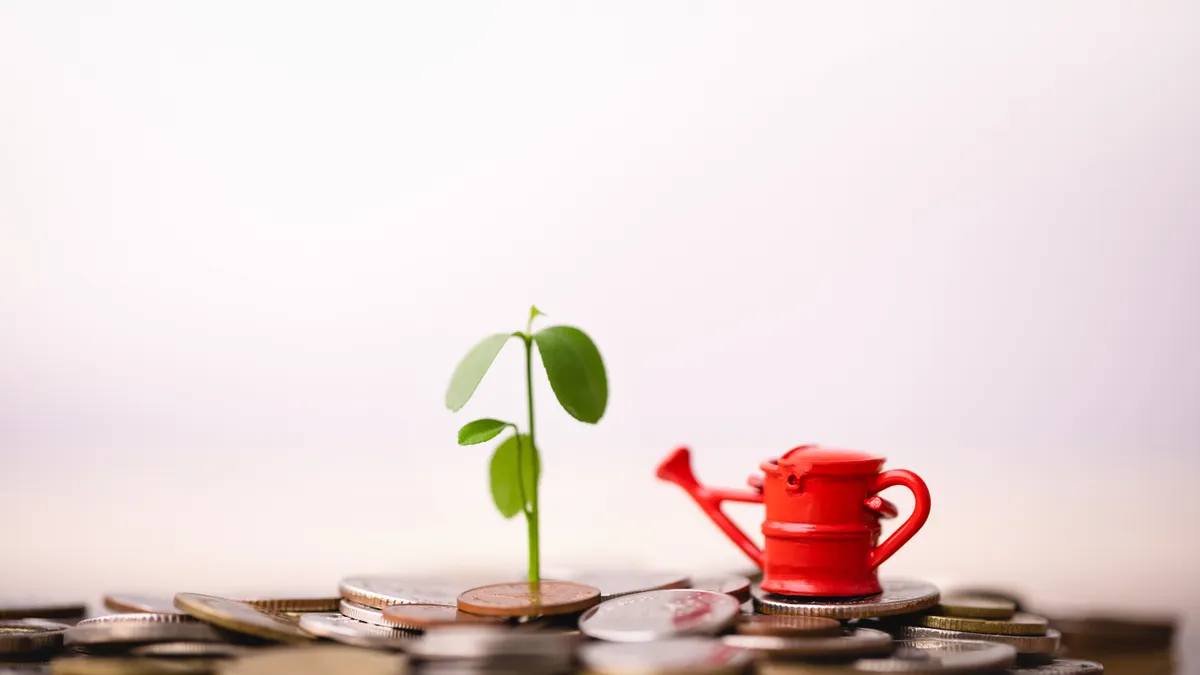 A watering can placed next to a sprouting plant coming from the ground, which is covered in coins.