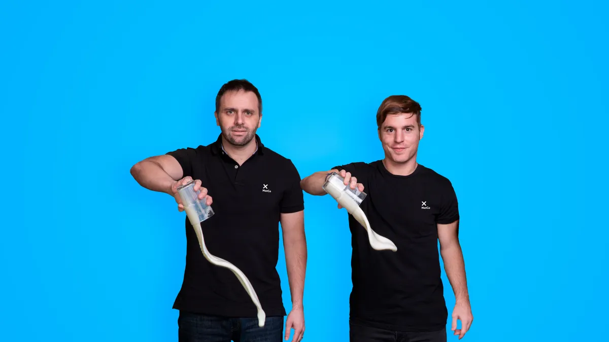 Karim Pichara and Matias Muchnick are dressed in black pants and shirts, pouring out glasses of milk on a blue background.