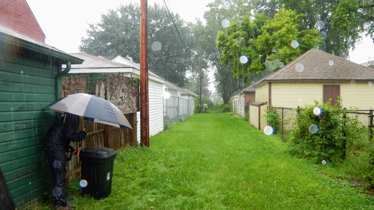 A person with an umbrella leans over a garbage bin in an alley as it rains.