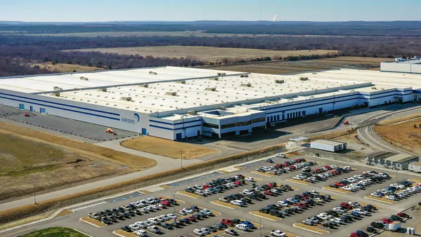 A sky view of a very large, one-story manufacturing plant, including a parking lot filled with cars, a road in front of the plant, and trees and fields beyond.