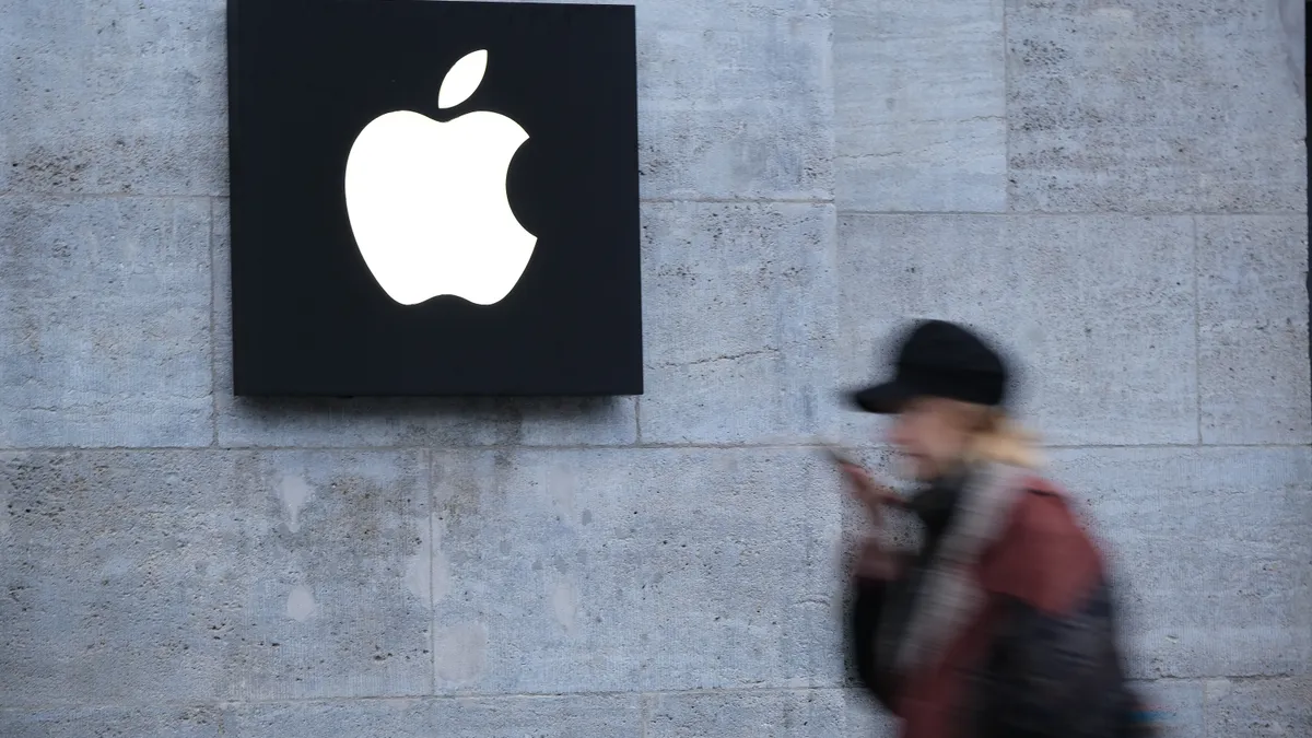 A woman holding a smartphone walks past a gray wall with an apple logo on a black sign.