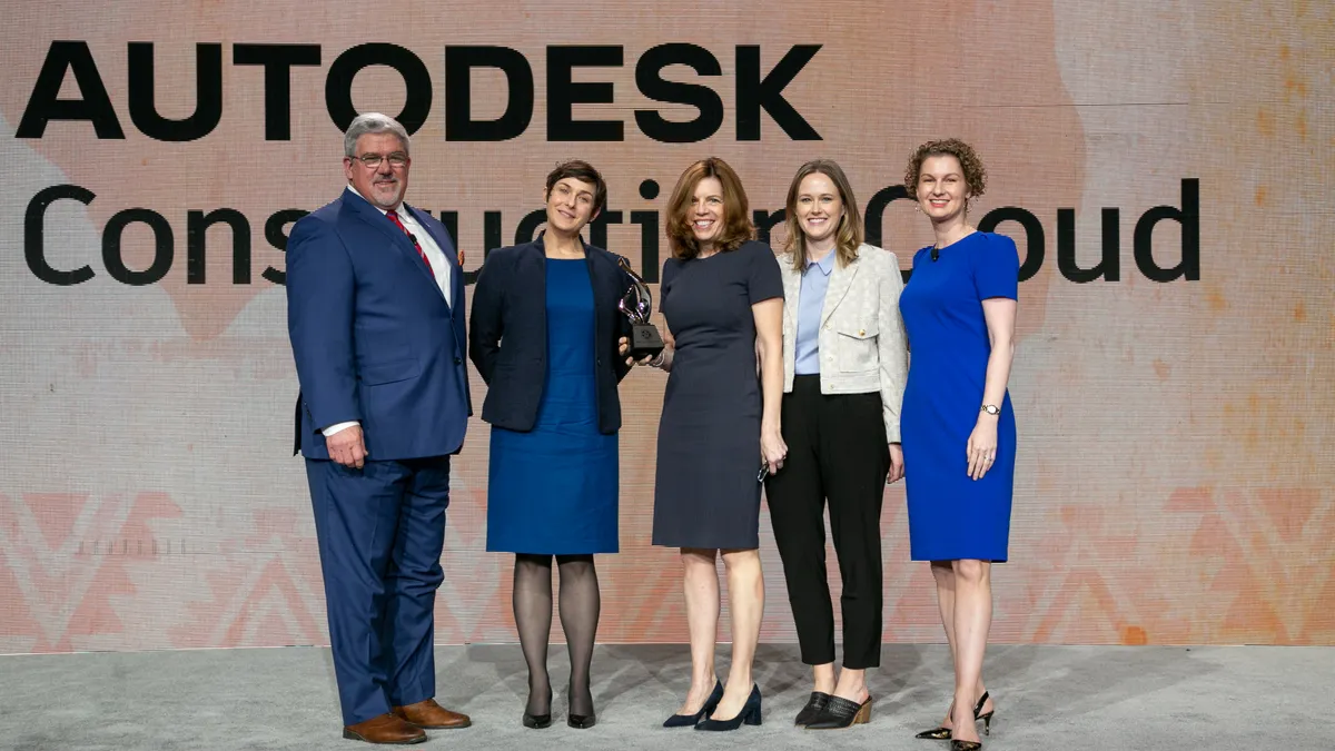 Five people stand on a stage with the centermost person holding an award. All are dressed formally, in suits and dresses. There are four women and one man, all smiling.