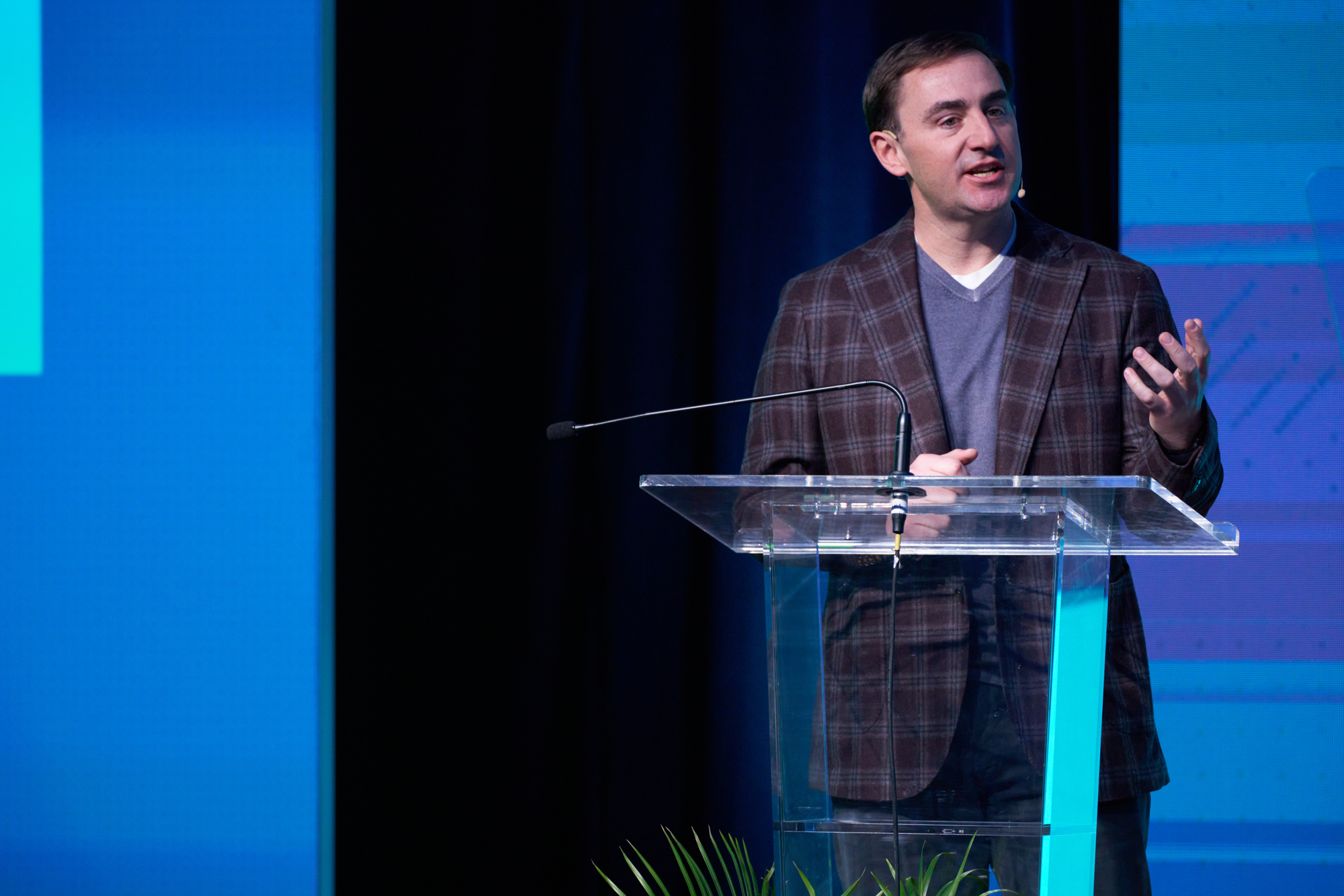 A person in a plaid jacket speaks at a podium, gesturing.