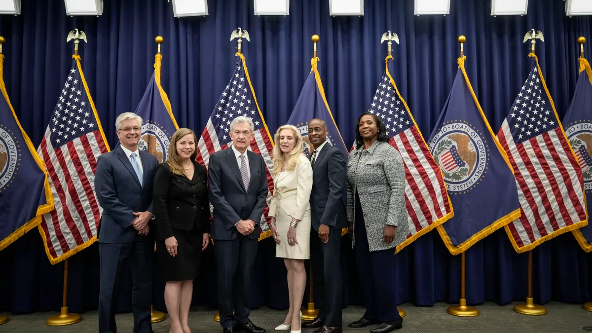 A group of six Federal Reserve Board members stand shoulder to shoulder with U.S. and state flags behind them.