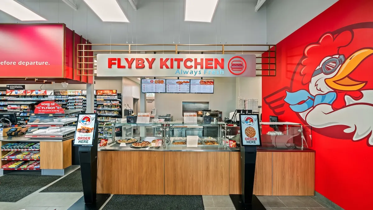 A photo of a foodservice counter in a store. A sign over the counter says "Flyby Kitchen. Always fresh." there are two ordering kiosks and several glass-covered display areas. A logo on the wall to the right shows a chicken in aviation goggles and a scarf.