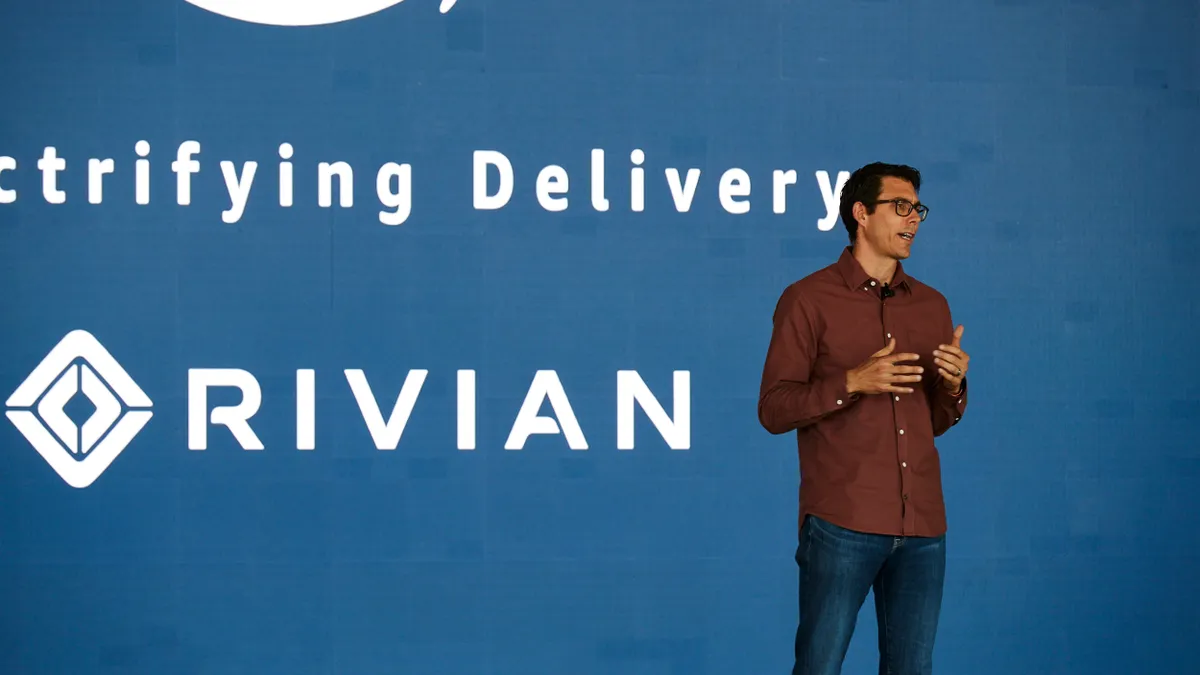 Rivian CEO RJ Scaringe speaks on the right side of a stage in front of a blue backdrop that says, "Electrifying Delivery" above the Rivian logo.