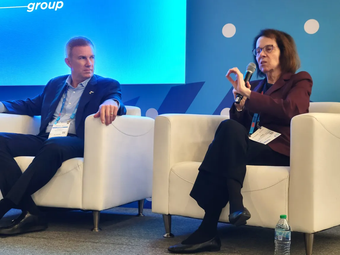 Two people sit in white chairs. The person on the right speaks while the person on the left listens.