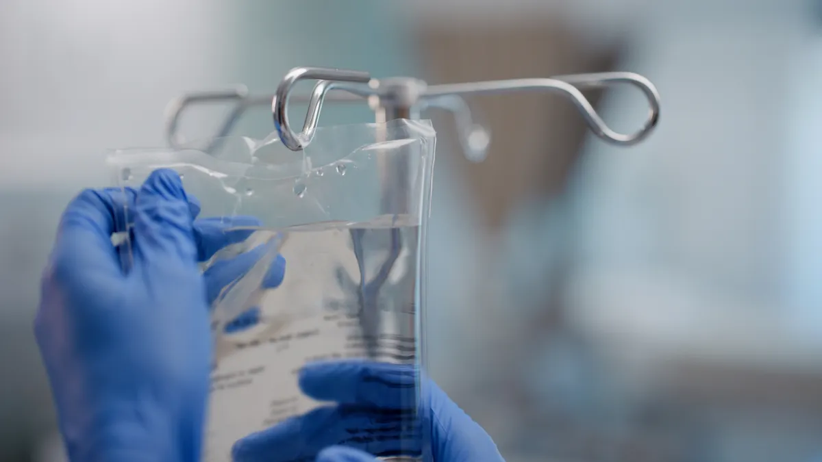 A person wearing gloves hangs a clear bag with fluid on a metal hook.