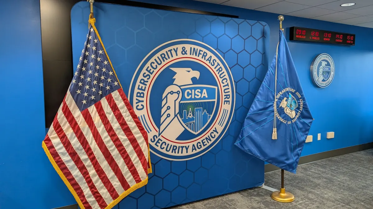 An American flag and a flag bearing the seal of the Cybersecurity and Infrastructure Security Agency (which features an eagle holding a shield with elements of a skyline on it) flank a large upright square panel bearing the same CISA seal. On the wall to the right of the panel and the flags, a row of digital clocks shows the time in the four major U.S. time zones.