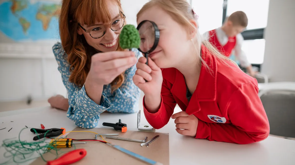 An adult and a student are sitting at a table. The student has a magnifying glass to their head and the teacher is holding an object in front the magnifying glass. of the