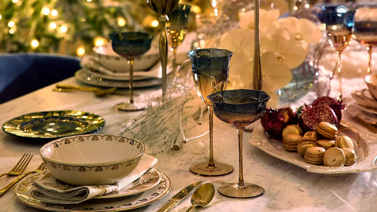 A table set with stemmed glasses, plates, bowls, platters and candlesticks with lit Christmas trees in the background.